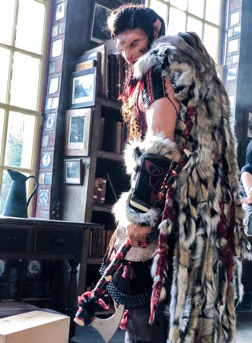 Man looking over his shoulder wearing cat like prosthetic makeup in a black, white, ivory and red Beast costume with beaded chest plate, celtic bracers and fur cloak.